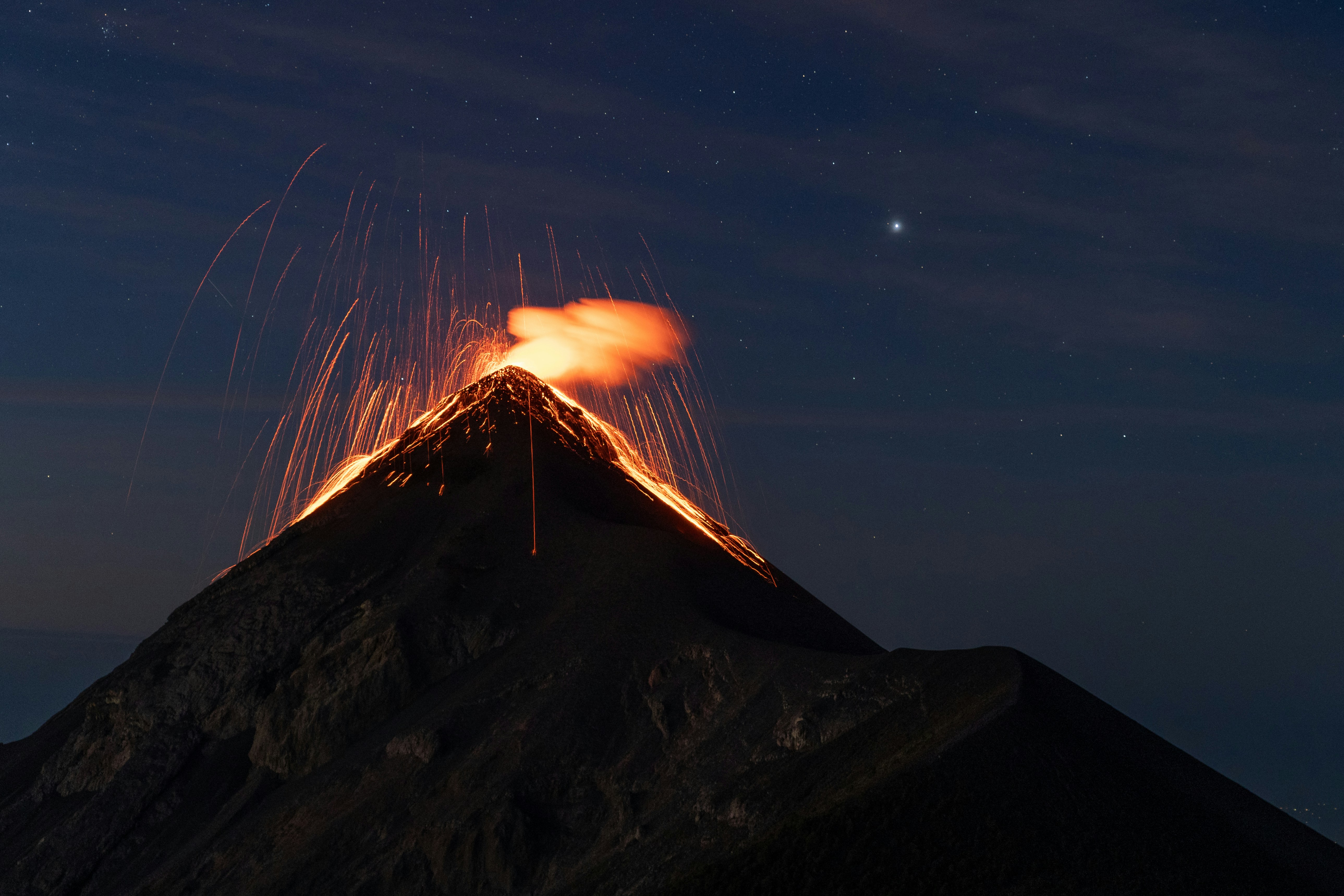 Mochileiros sentados numa crista escura a observar uma erupção vulcânica laranja brilhante contra um céu estrelado